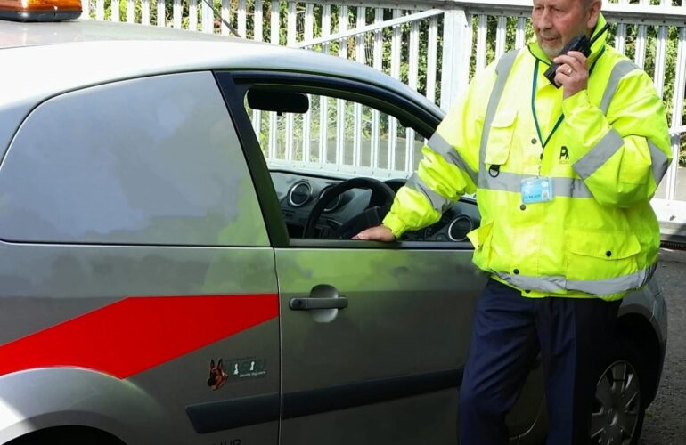 Security officer in a high-visibility jacket using a radio beside a marked patrol vehicle